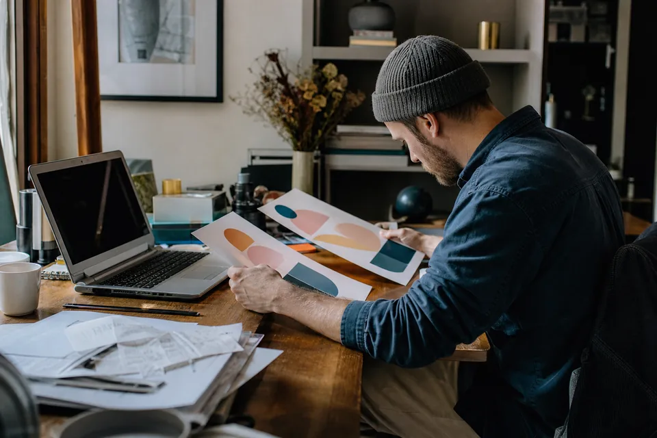 Self-employed designer comparing two printed price sheets at a desk with a laptop and a stack of 1099 forms