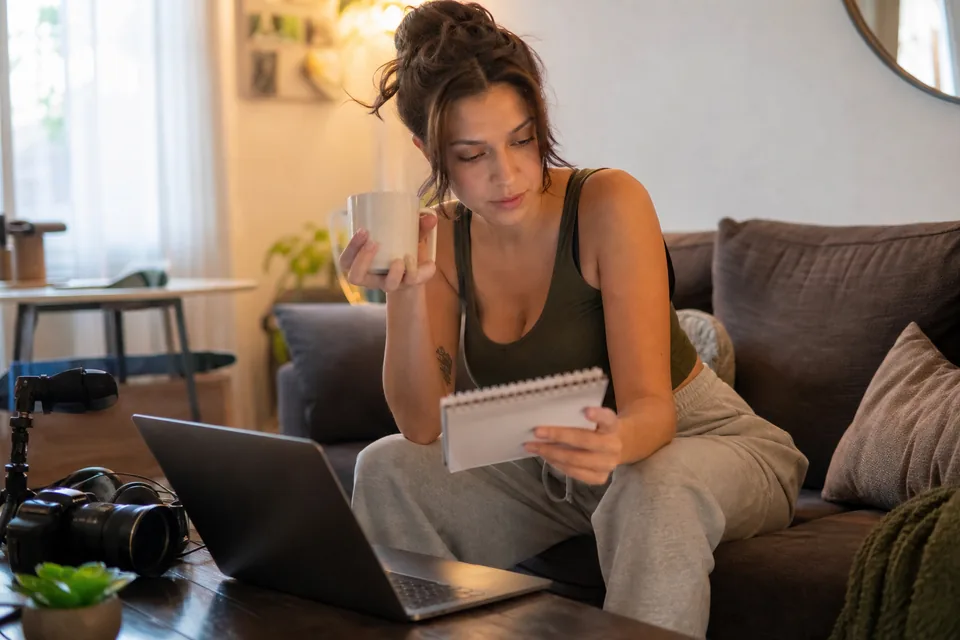 Creator on a couch comparing numbers on a notepad beside a laptop and camera