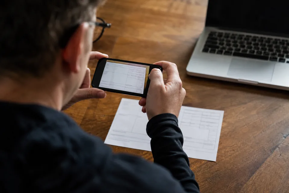 Hands photographing a paper W-2 with a smartphone on a wooden table to upload it for filing